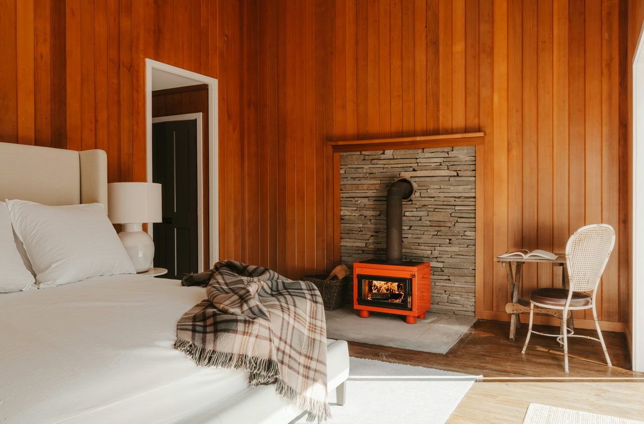 Hotelzimmer mit Holzvertäfelung, orangem Kaminofen, Steinwand, Leinenbett und Wolldecke