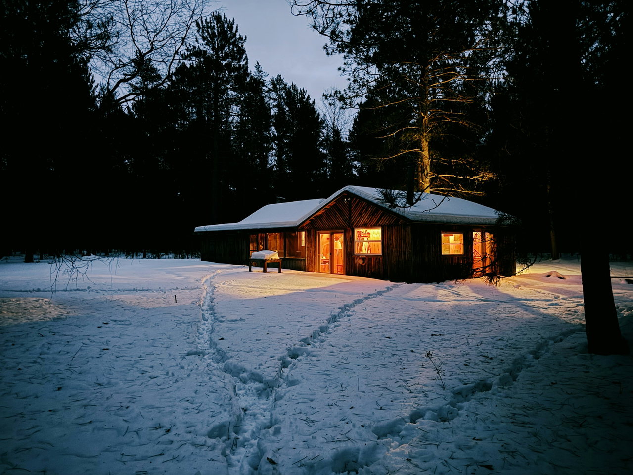 Holzhütte mit warm erleuchteten Fenstern im Schnee, Fußspuren im Vordergrund, Winterdämmerung
