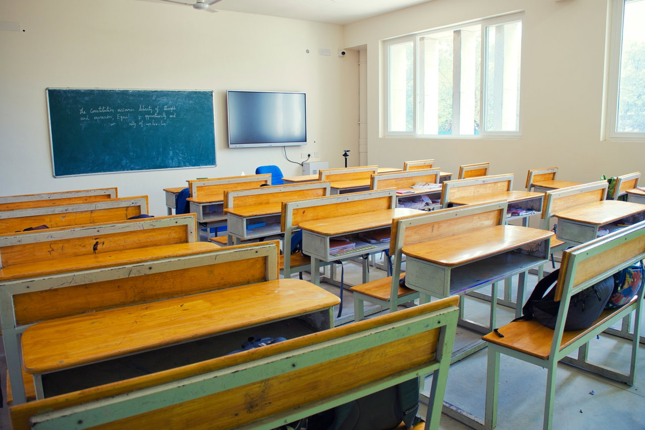 Heller, leerer Klassenraum mit Holztischen, Fenstern, Tafel und Bildschirm an der Stirnwand