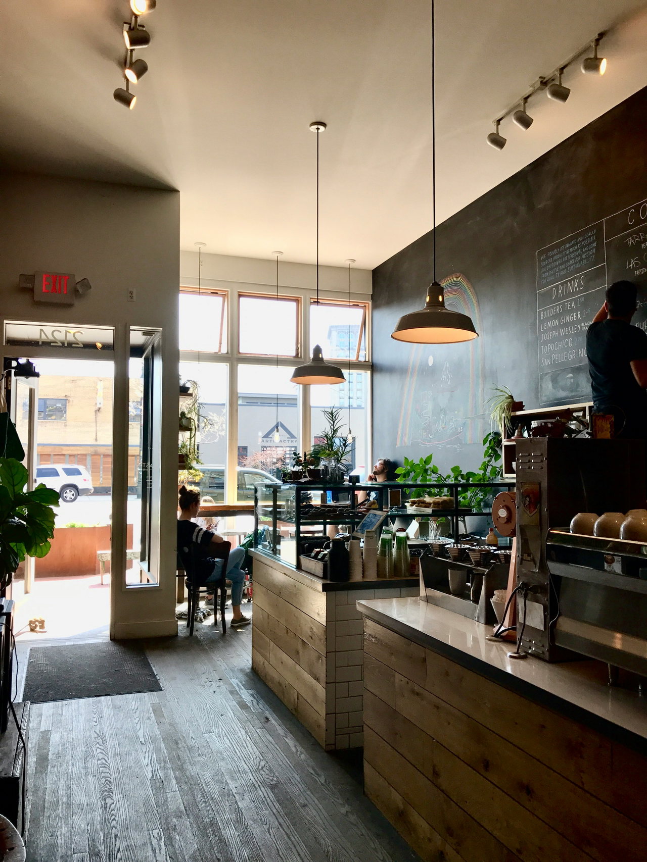 Café-Innenraum mit Holztresen, Espressomaschine, Kreidetafel, Hängeleuchten und Pflanzen am Fenster