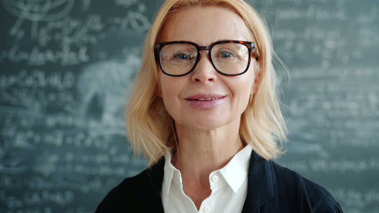 Porträt einer blonden Frau mit Brille und dunkler Strickjacke vor einer Schiefertafel mit Formeln, freundlicher Blick in die Kamera