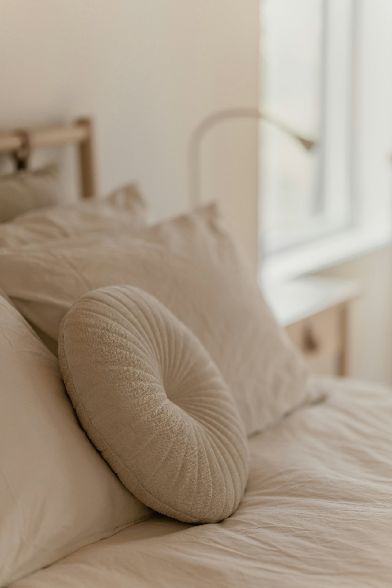Bett mit cremefarbener Leinenbettwäsche, Kissen und rundem Zierkissen im weichen Licht am Fenster