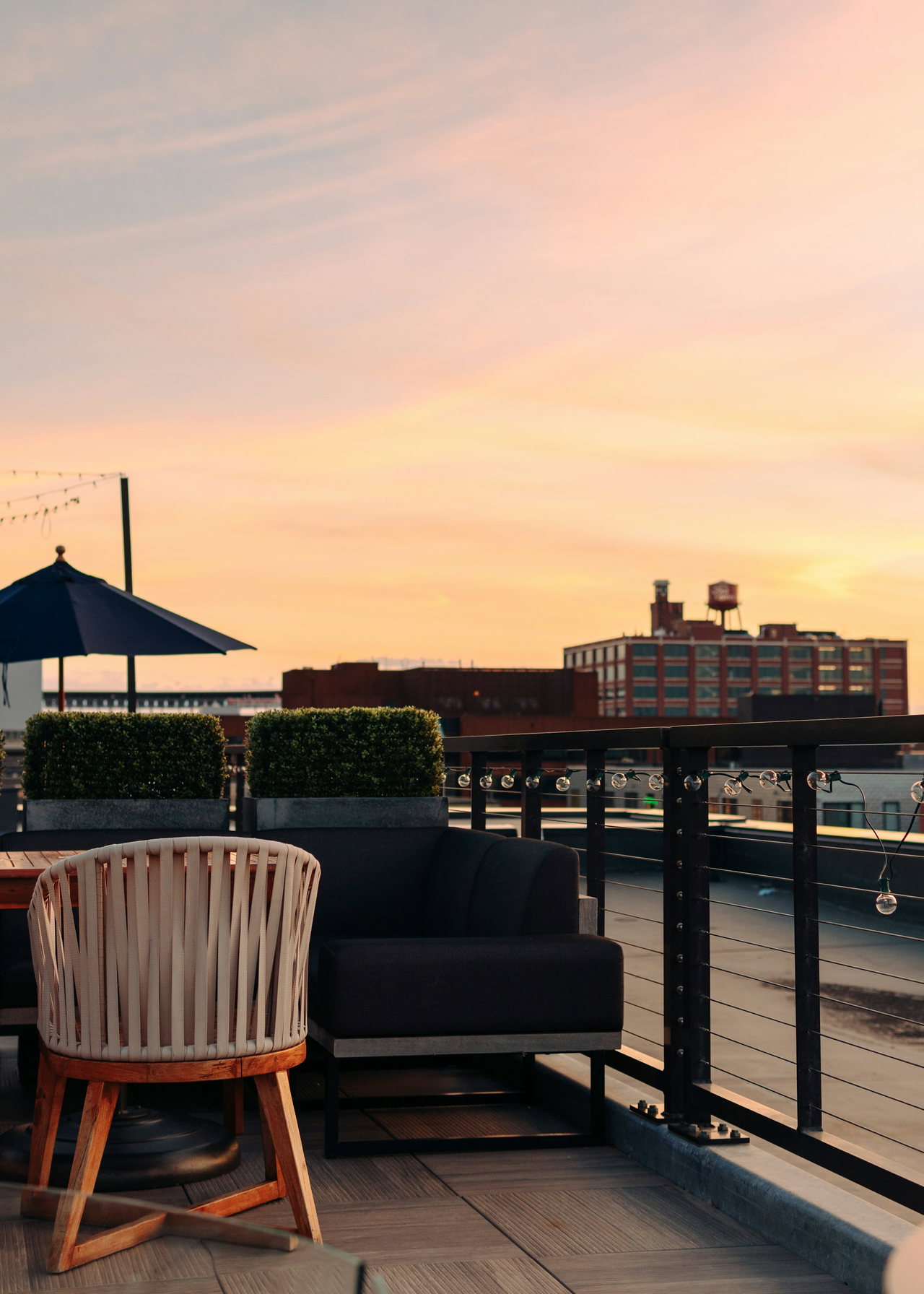 Dachterrasse mit geflochtenem Sessel, dunklem Loungesofa, Sonnenschirm und Lichterkette unter rosafarbenem Abendhimmel, dahinter alte Backsteinbauten.