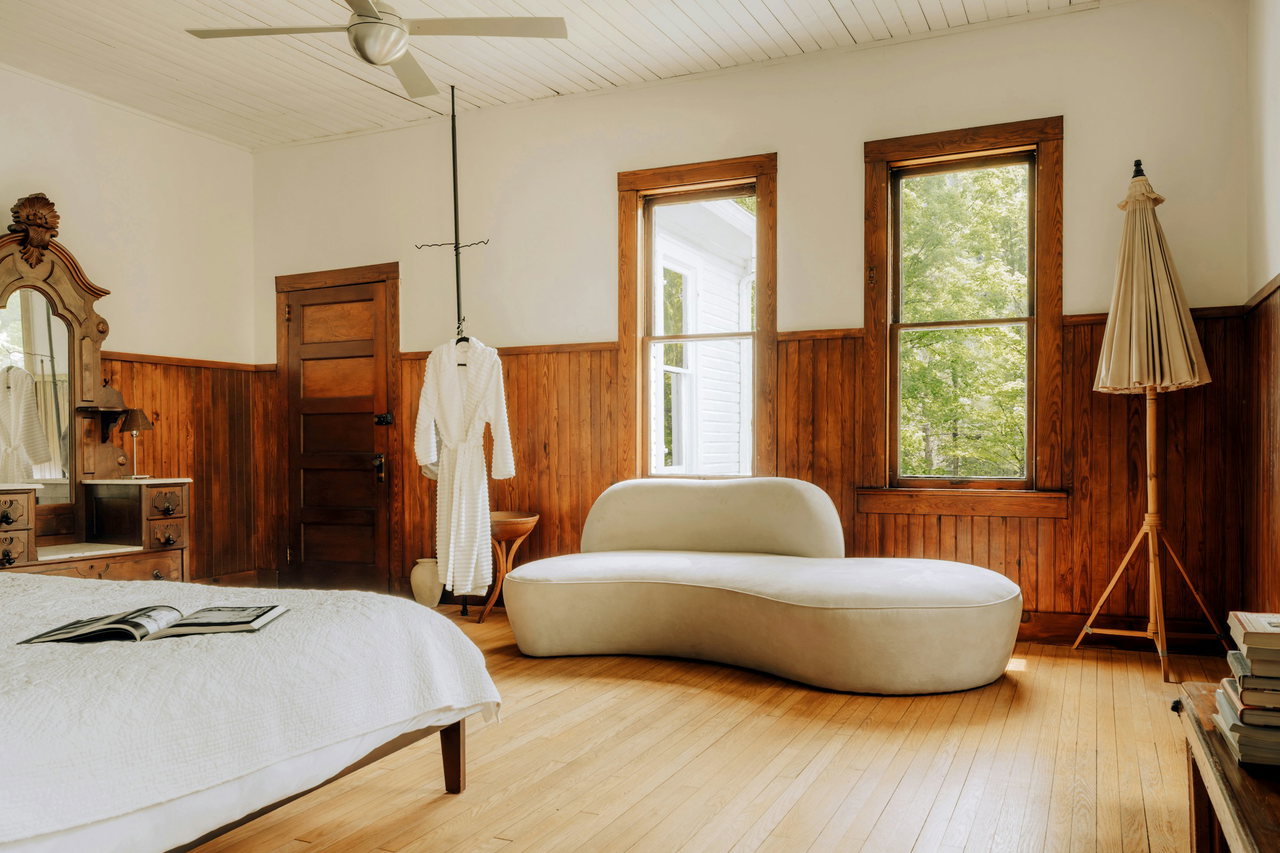 Helles Zimmer mit Holzvertäfelung, Holzdielen, weißem Bett, geschwungenem Sofa und Bademantel am Ständer neben Fenstern