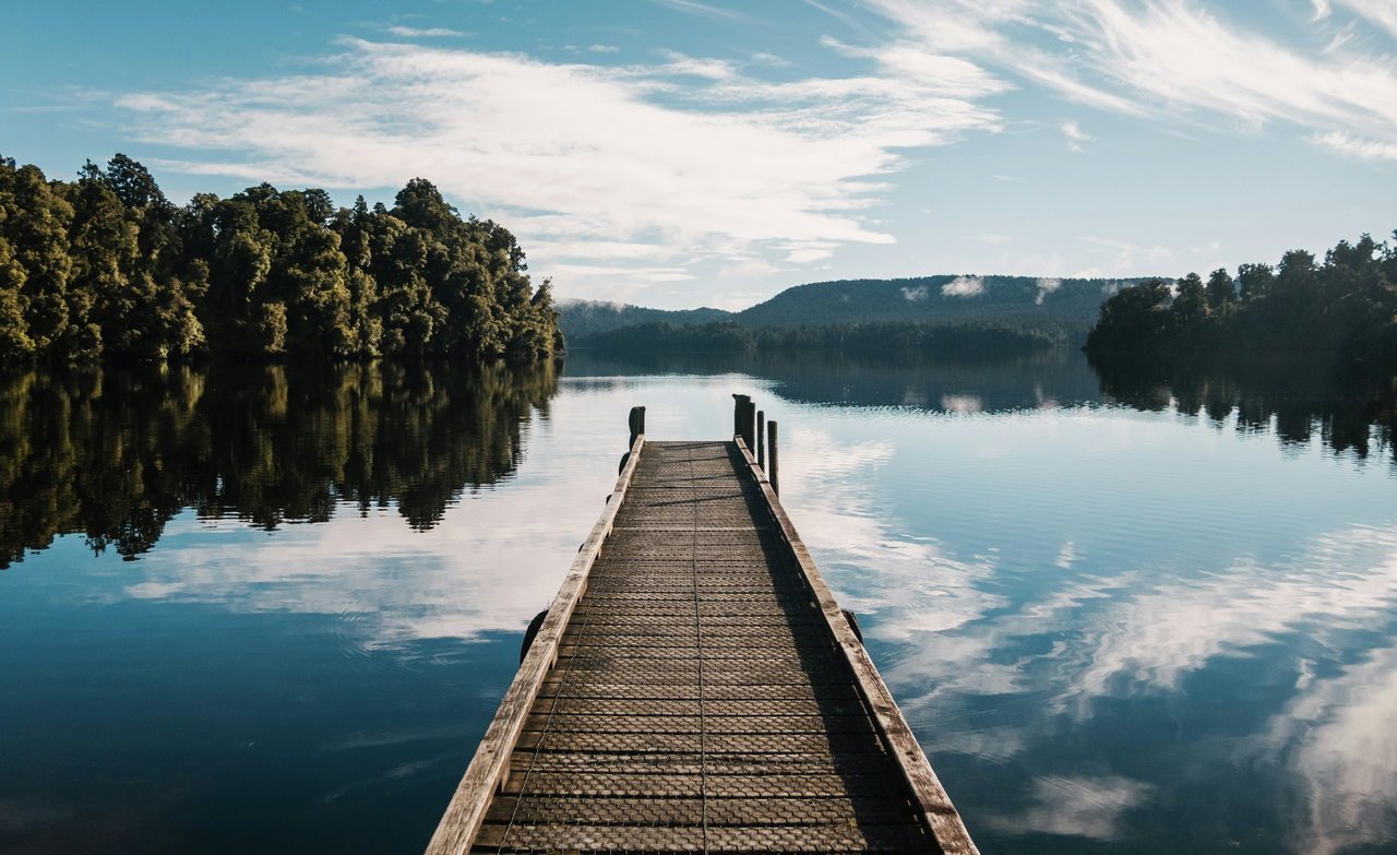 Holzsteg führt auf einen stillen See, in dem sich Wald und Wolken spiegeln.