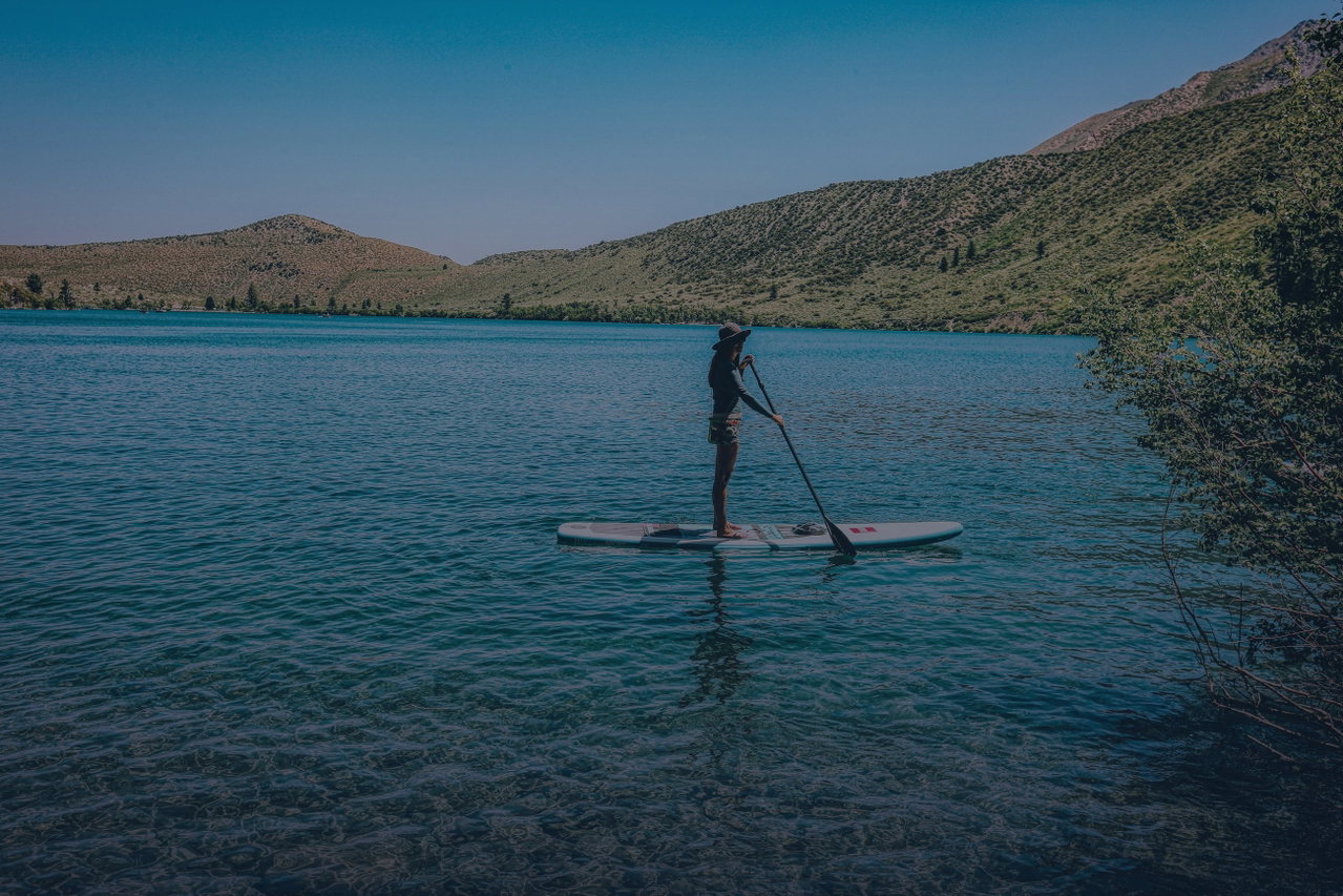 Eine Person steht auf einem Stand-up-Paddleboard und paddelt über einen türkisfarbenen See vor bewaldeten Hügeln