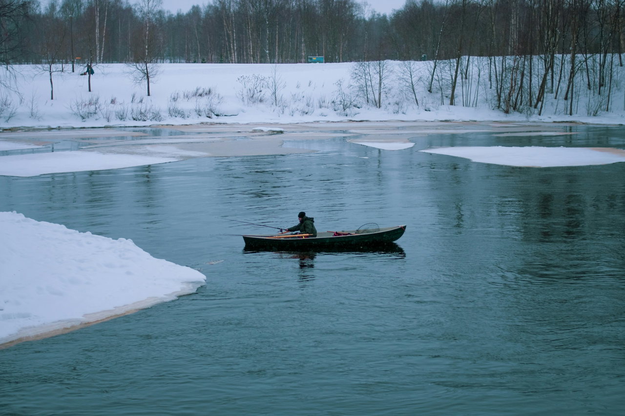 Ruderboot mit einer Person auf winterlich ruhigem Wasser, Eisschollen und verschneites Ufer im Hintergrund