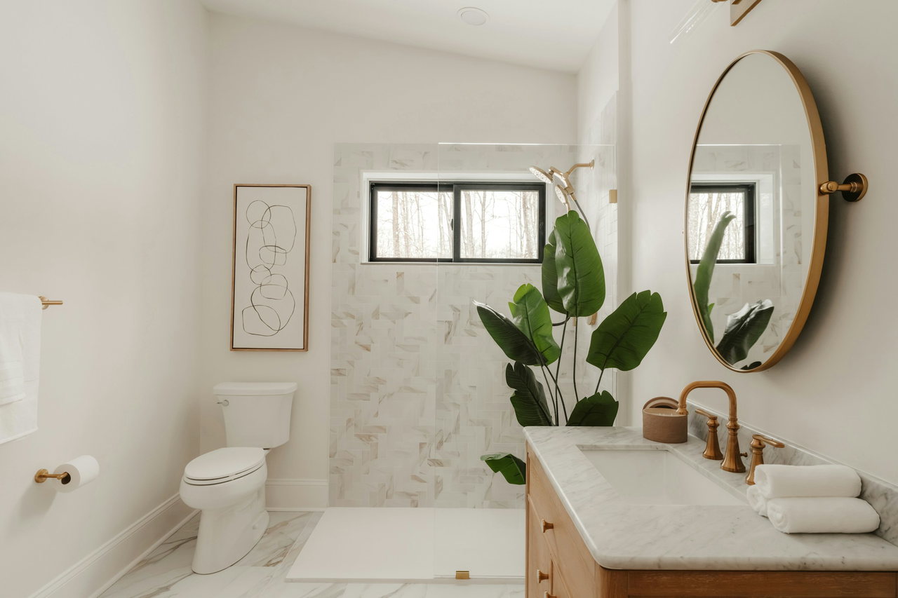 Helles Bad mit bodengleicher Dusche hinter Glas, rundem Spiegel im Messingrahmen und Waschtisch mit Marmorplatte, Holzunterschrank und Messingarmaturen.