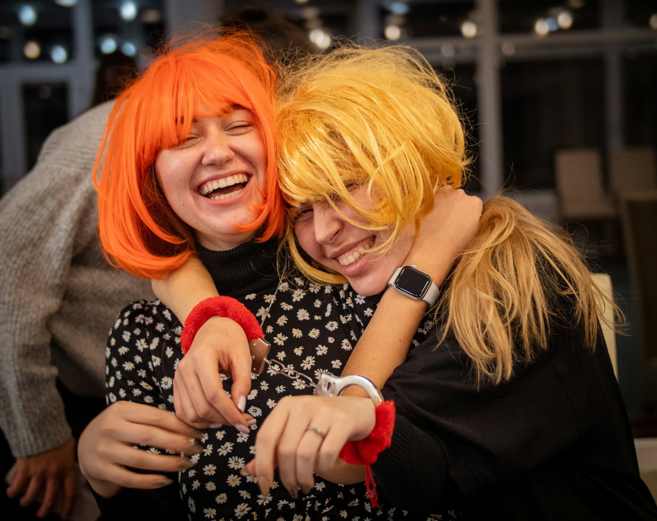Zwei lachende junge Frauen mit orangefarbener und blonder Perücke umarmen sich bei einer Verkleidungsparty