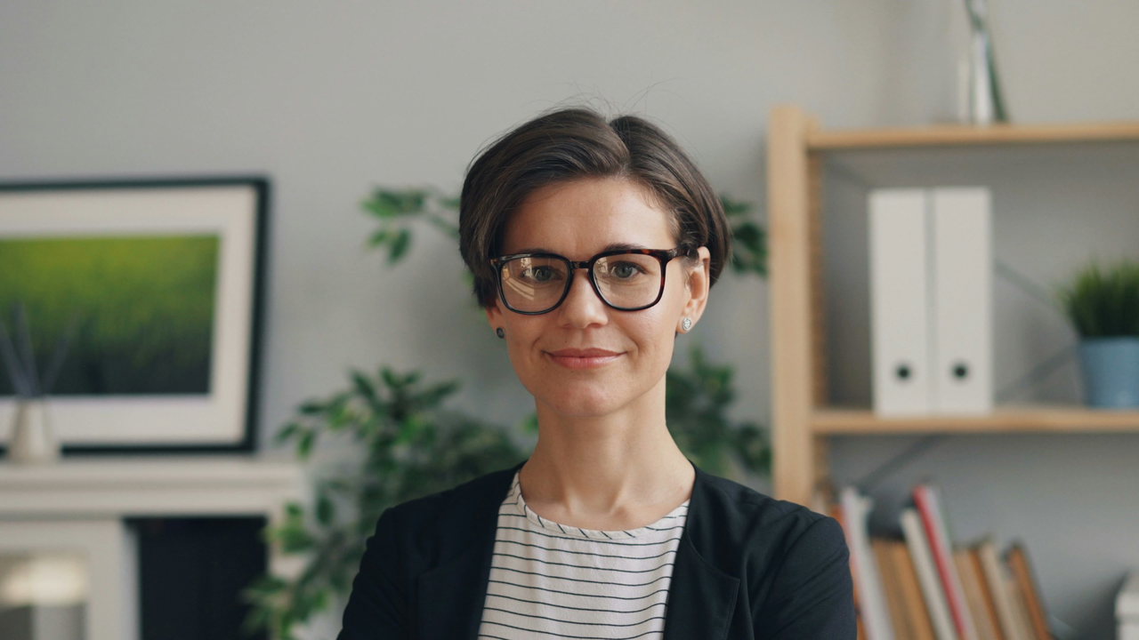 Porträt einer Frau mit dunklem Kurzhaarschnitt und Brille, schwarzer Blazer, gestreiftes Shirt, heller Raum mit Pflanzen