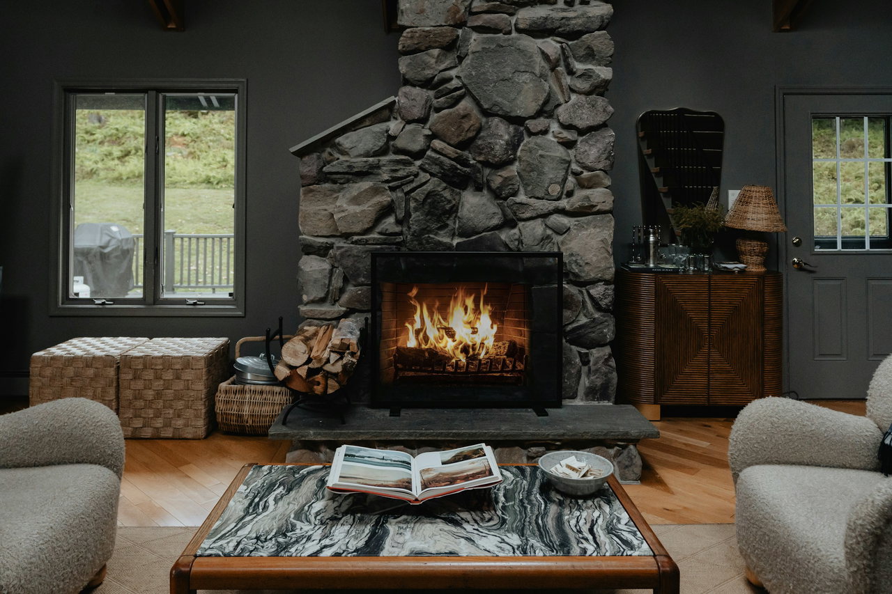 Kaminlounge mit loderndem Feuer im Natursteinkamin, zwei hellen Bouclé-Sesseln, Marmortisch mit Bildband und Brennholz daneben