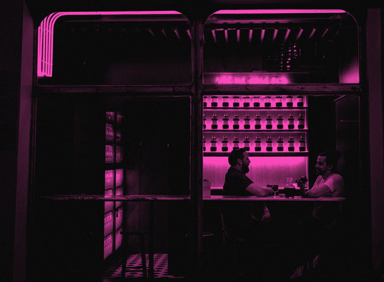 Symbolfoto: Neonbeleuchteter Bartresen mit zwei Gästen von hinten, Schwarzweiß mit Magenta-Duplex