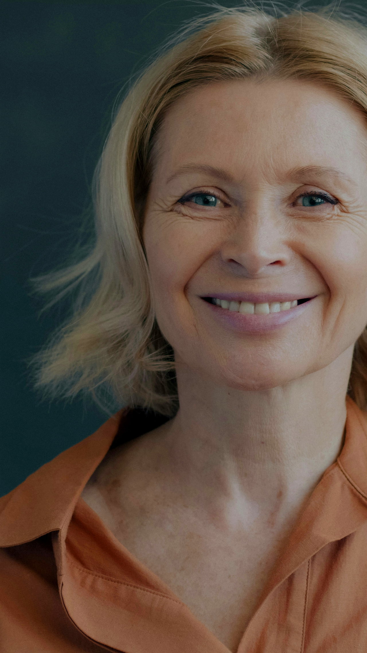 Porträt einer lächelnden Frau mit blonden Haaren und orangefarbener Bluse vor dunklem Hintergrund