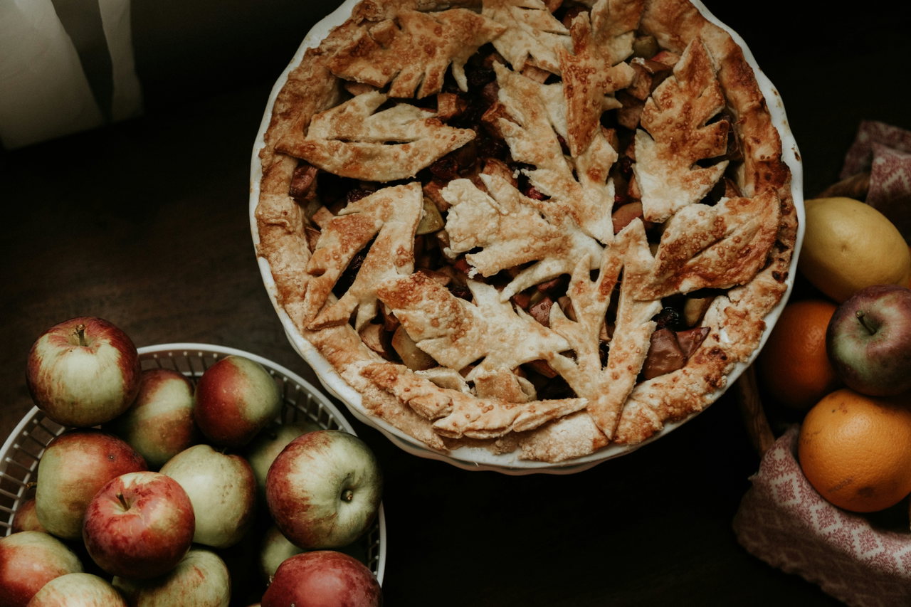 Gedeckter Apfelkuchen mit Teigblättern auf einem dunklen Tisch, daneben Schalen mit roten und grünen Äpfeln