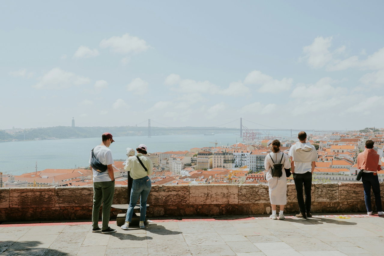 Mehrere Menschen stehen an einer Mauer und blicken über die roten Dächer Lissabons zum Tejo und zur Brücke