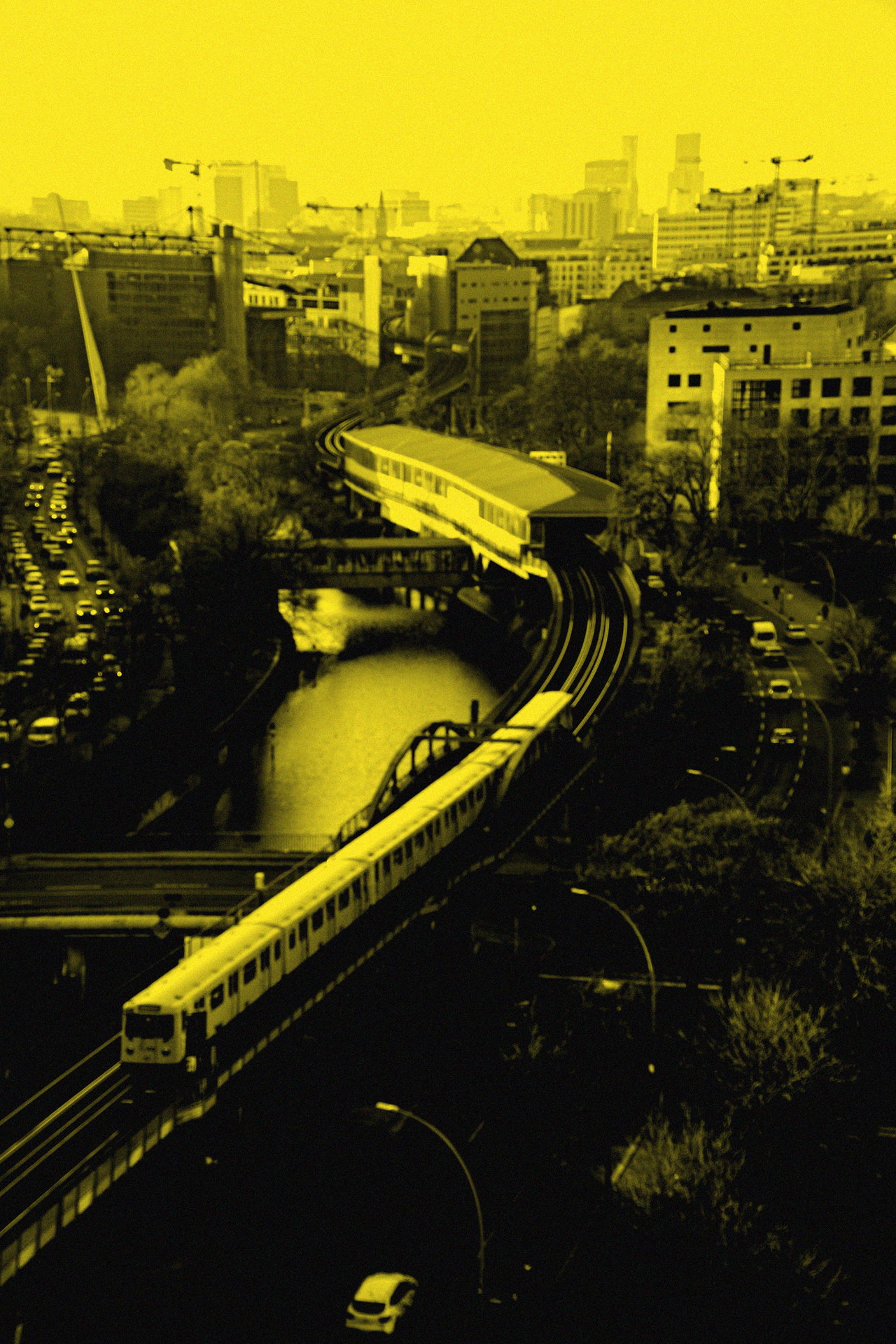 Hochbahnzug auf der Kurve über dem Kanal, Stadtsilhouette dahinter, Gelb-Duplex