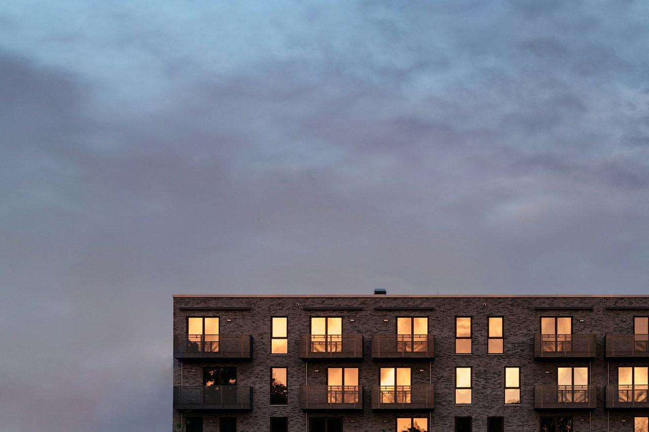 Obere Geschosse eines Wohnhauses aus dunklem Klinker mit Balkonen in der Abenddämmerung; in den Fenstern spiegelt sich warmes Abendlicht, darüber ein weiter, wolkiger Himmel.