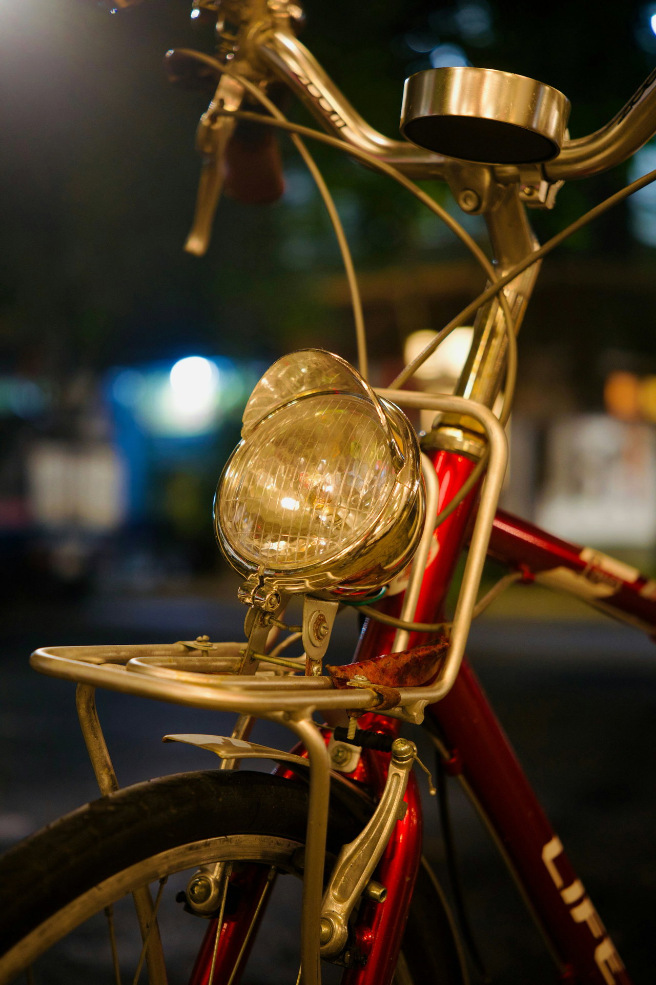 Goldener Retro-Scheinwerfer, Klingel und Gepäckträger an einem roten Fahrrad, abends vor dunklem, unscharfem Hintergrund