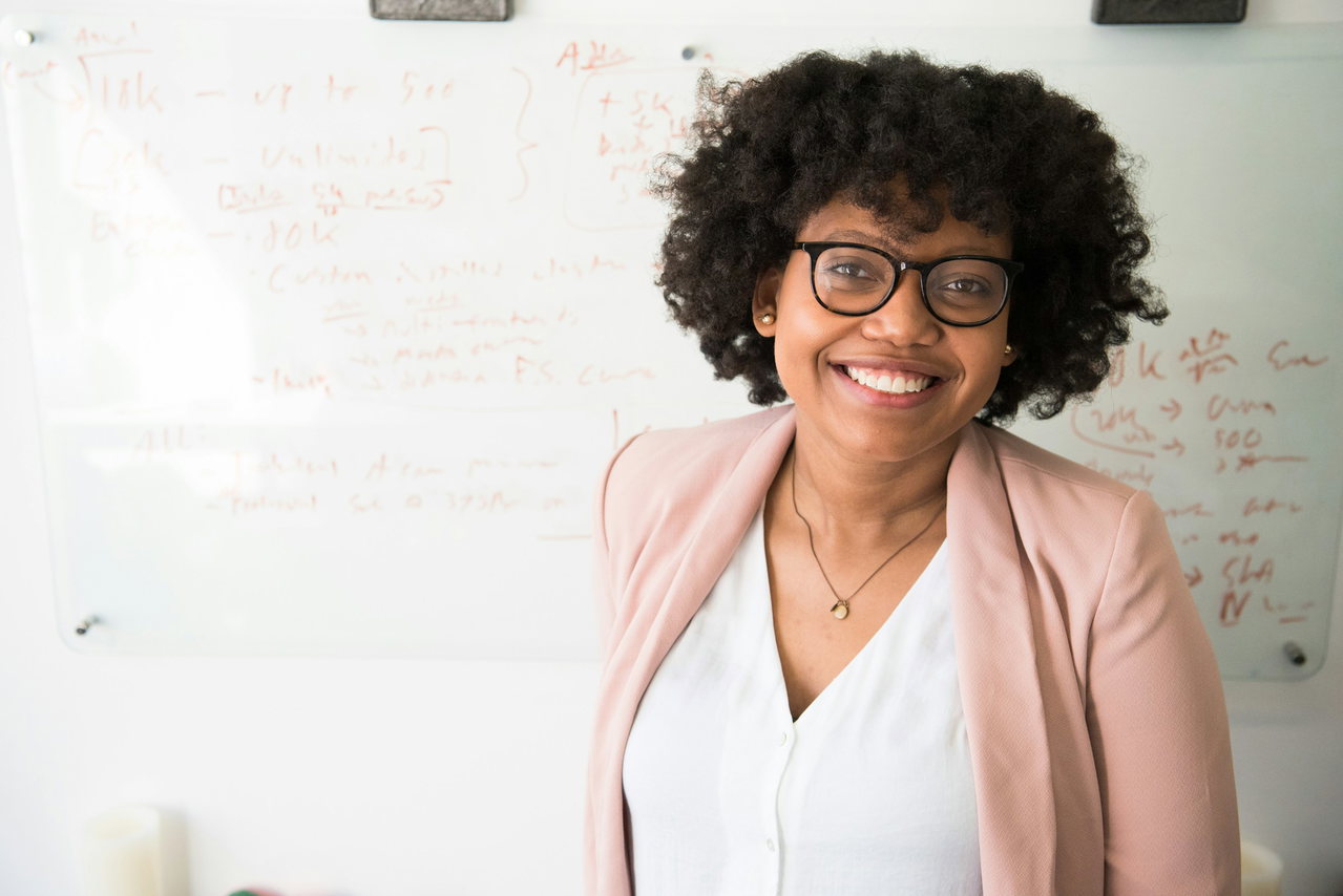 Lächelnde Frau mit lockigem Afro, Brille und rosa Blazer steht vor einem Whiteboard