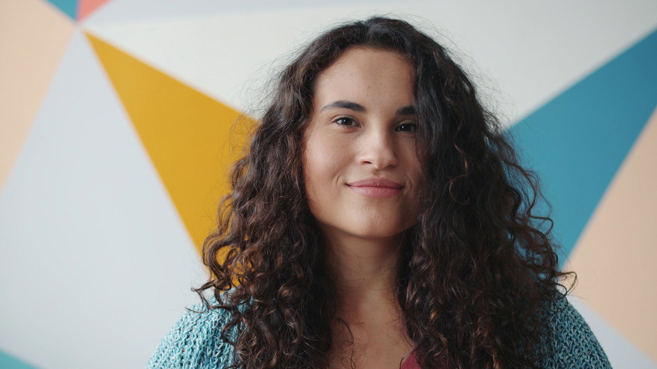 Junge Frau mit langen dunklen Locken blickt ruhig lächelnd in die Kamera vor einer hellen Wand mit bunten Farbflächen