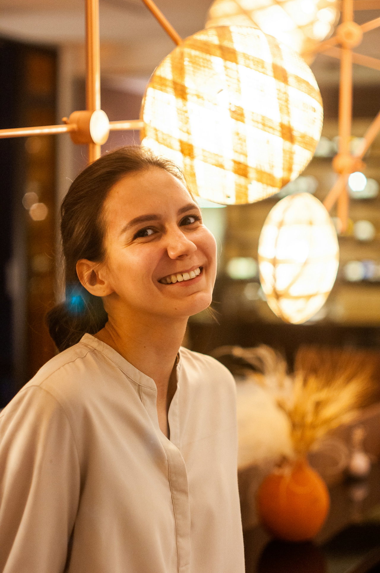 Porträt einer jungen Frau in heller Bluse, die lächelnd in die Kamera blickt, warme Lampen im Hintergrund