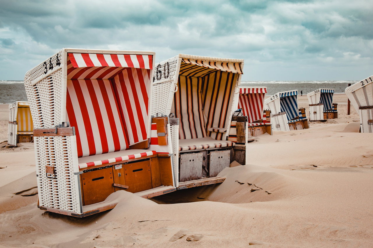 Bunt gestreifte Strandkörbe aus weißem Korbgeflecht stehen mit Nummern im Sand, dahinter die See unter Wolken