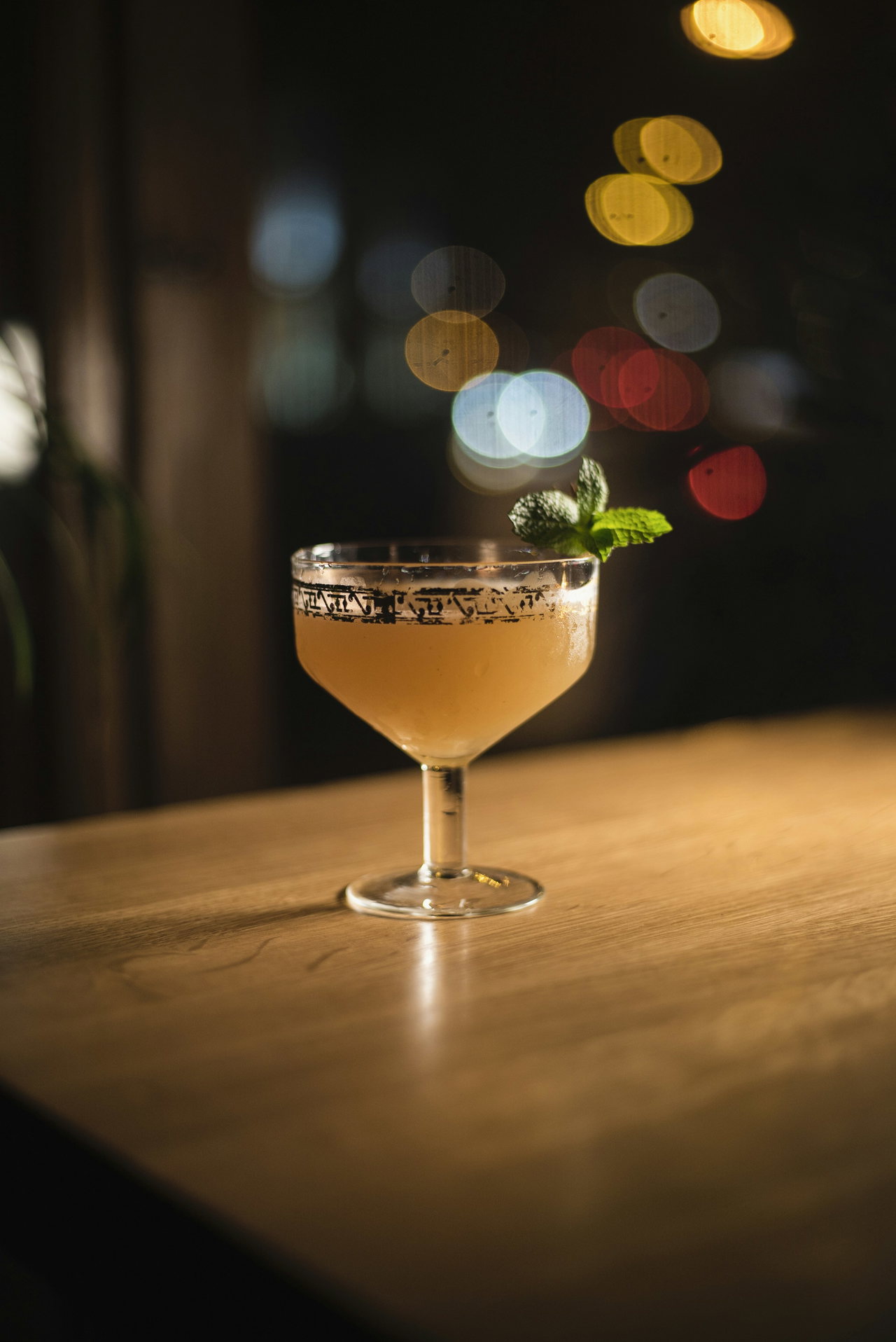 Goldfarbener Cocktail mit Minzzweig in einer Cocktailschale auf dem Holztresen, im Hintergrund warme Lichtpunkte