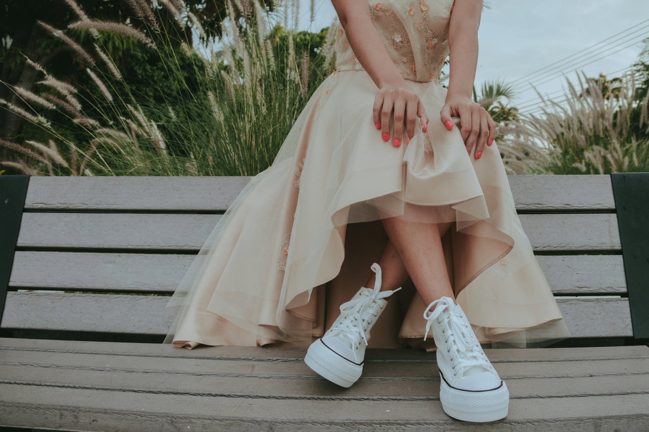 Weiße Sneaker unter einem cremefarbenen Abendkleid, die Trägerin sitzt auf einer Holzbank