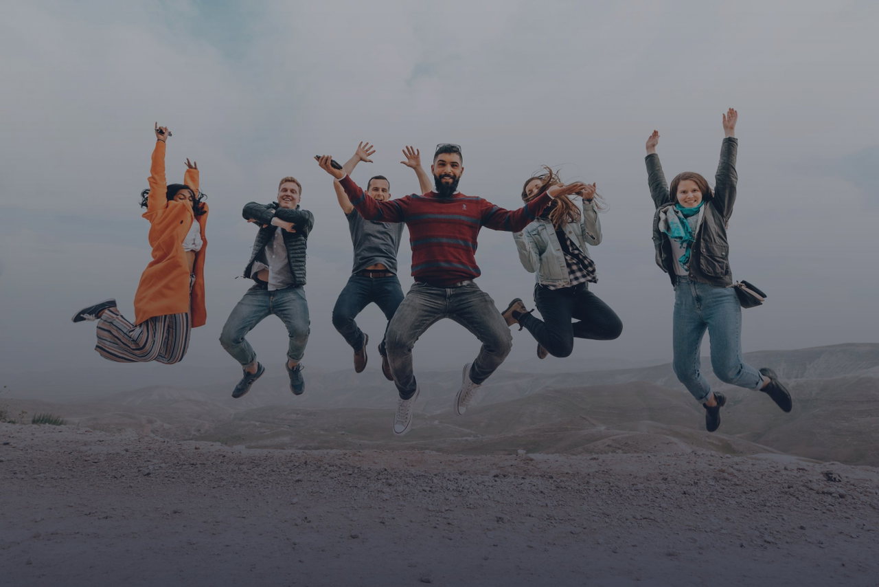 Sechs junge Leute springen gemeinsam jubelnd in die Luft, vor hellem Himmel und weiter Landschaft