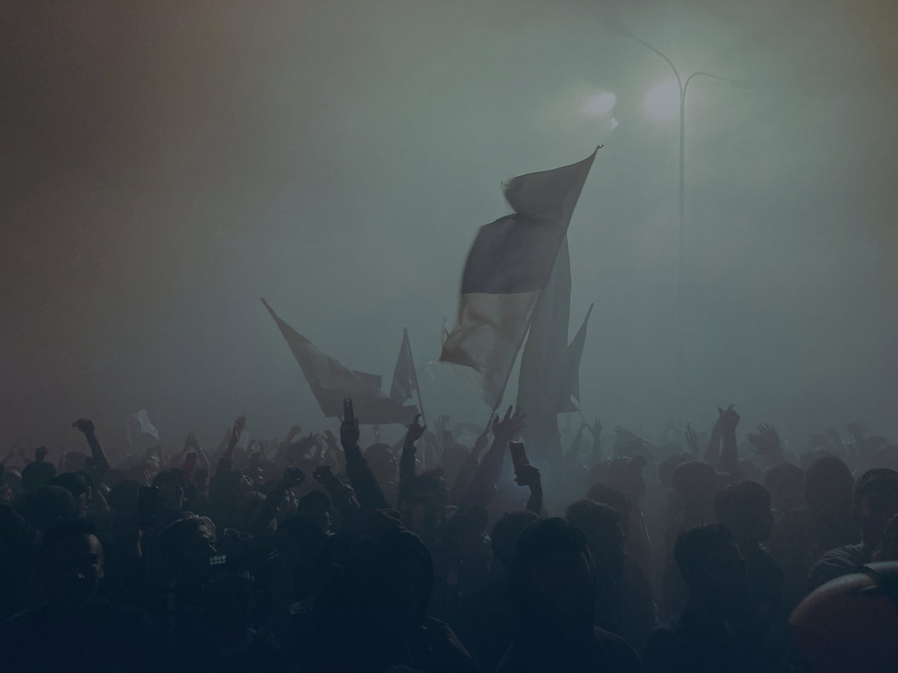 Fahnen wehen im Nebel und Flutlicht über den Köpfen der Fans in der Kurve, Blick von hinten