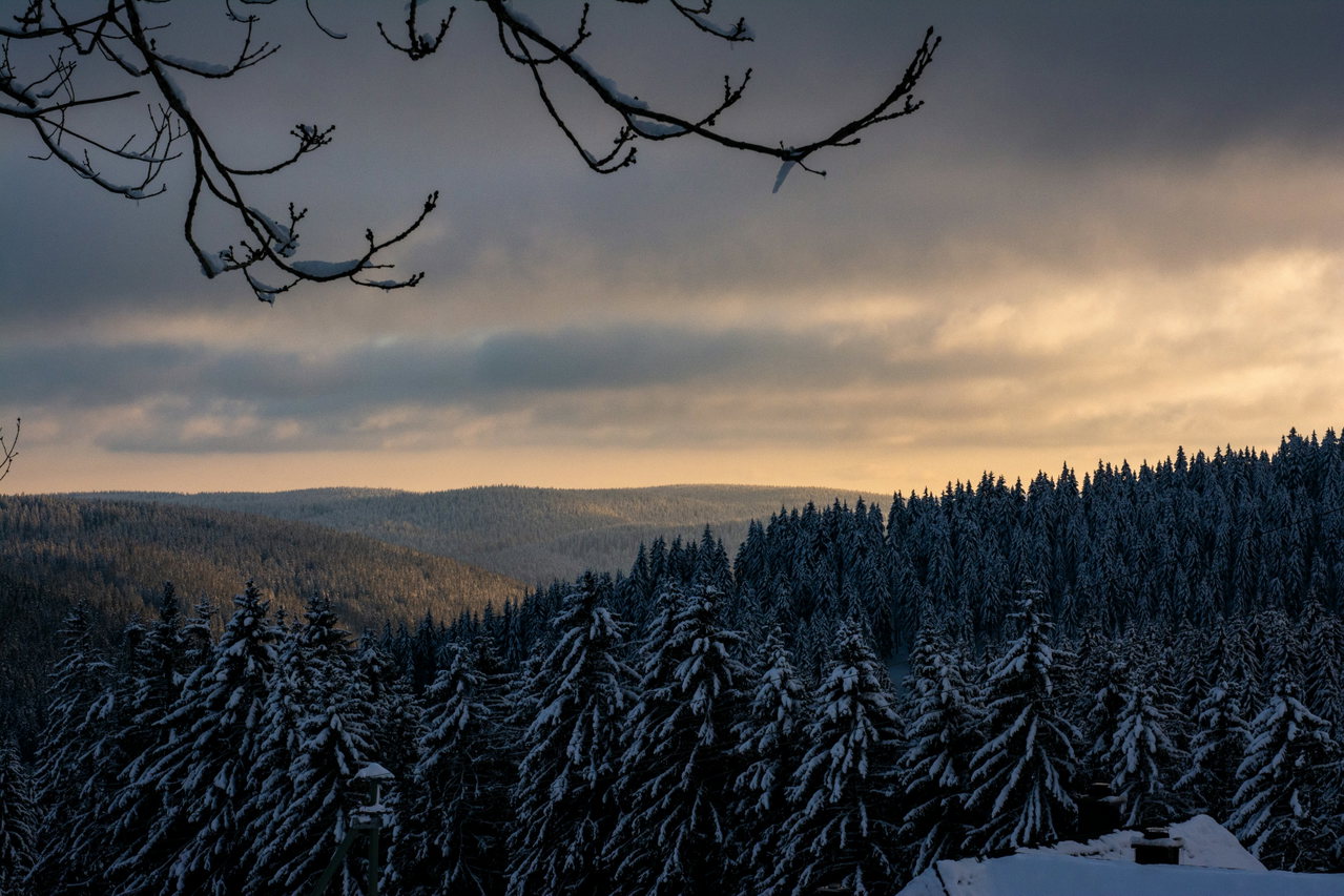 Weiter Blick über bewaldete, verschneite Mittelgebirgshügel im goldenen Abendlicht unter dunklen Wolken