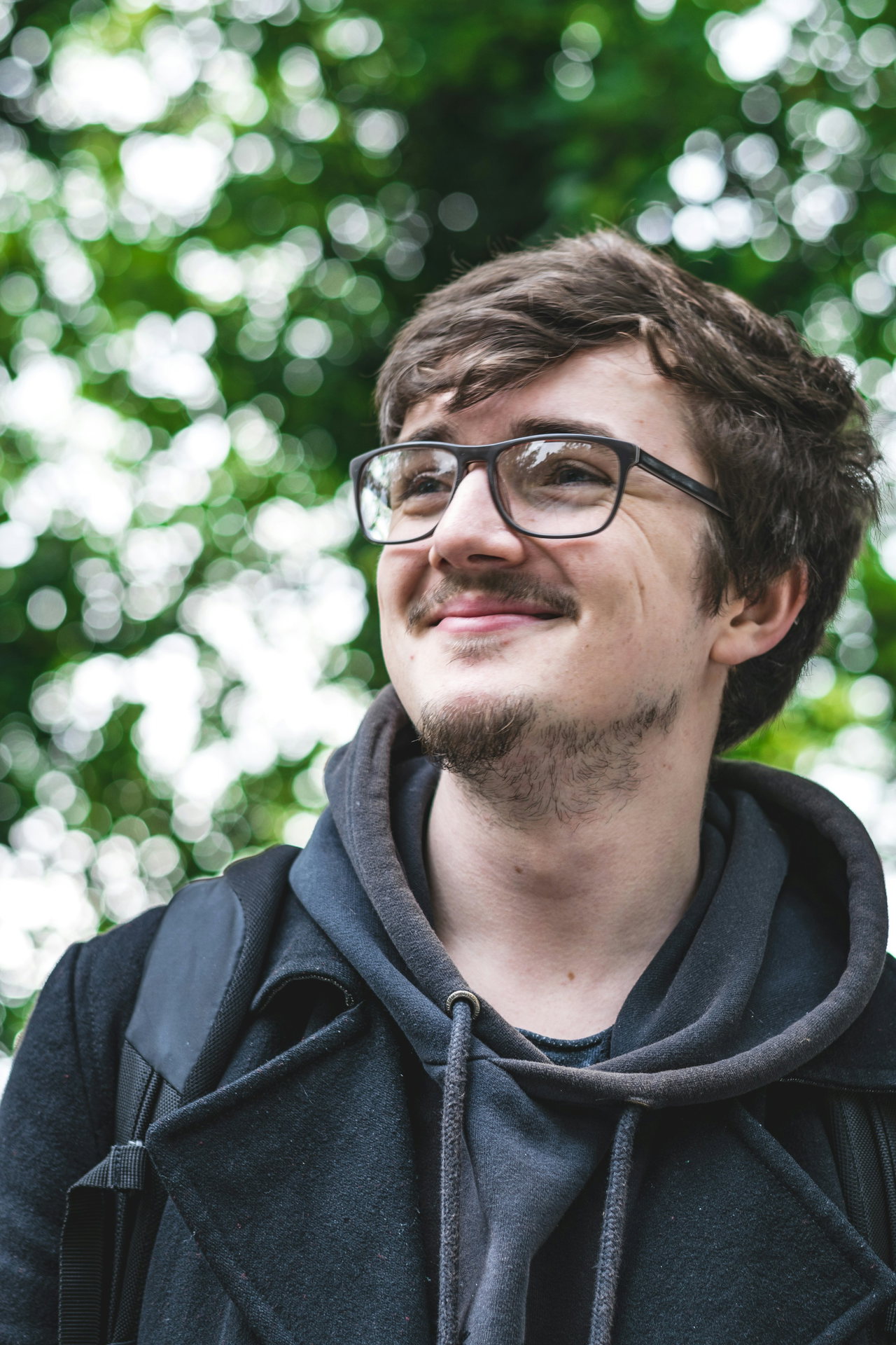 Junger Mann mit Brille und Schnurrbart in dunklem Kapuzenpullover blickt lächelnd zur Seite, grüne Bäume unscharf dahinter