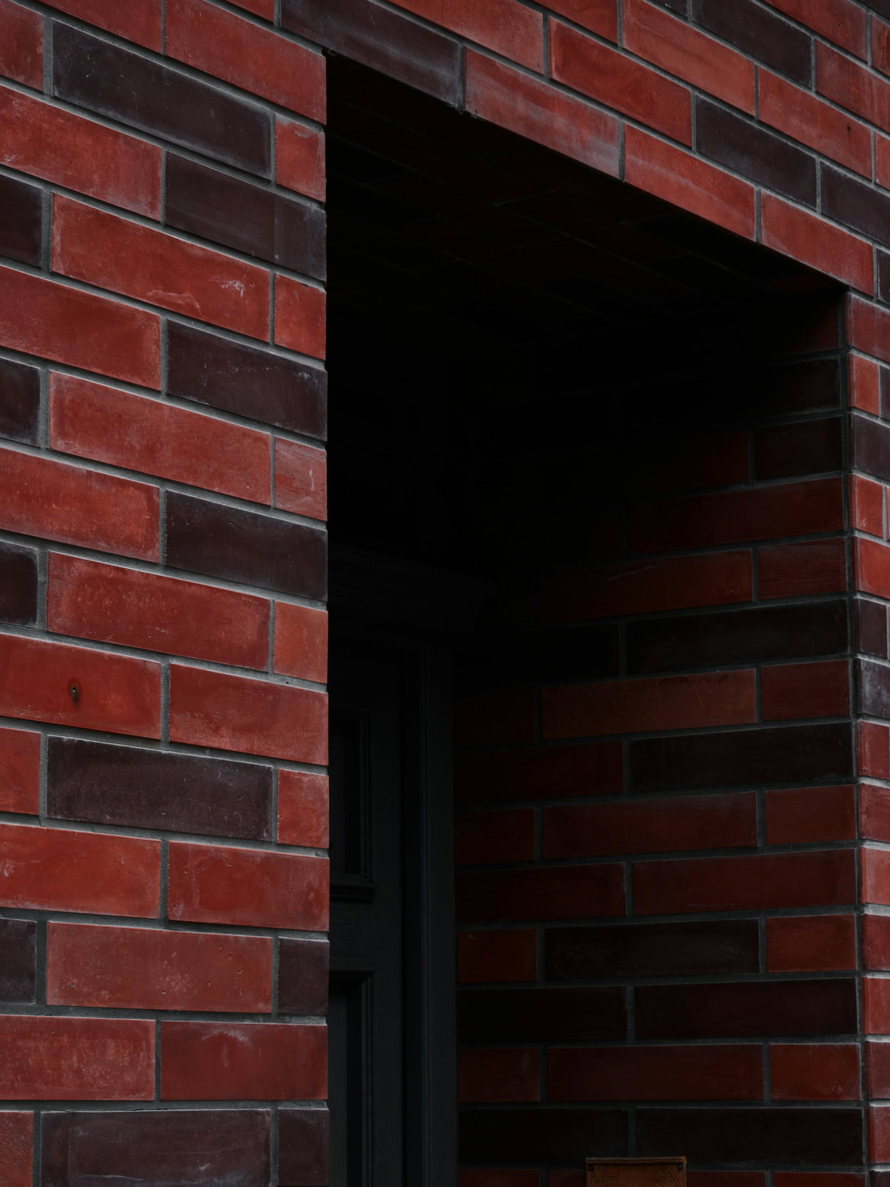 Tiefer, schattiger Eingang in einer Fassade aus roten und dunkelvioletten Klinkern; auch die Laibung ist gemauert.