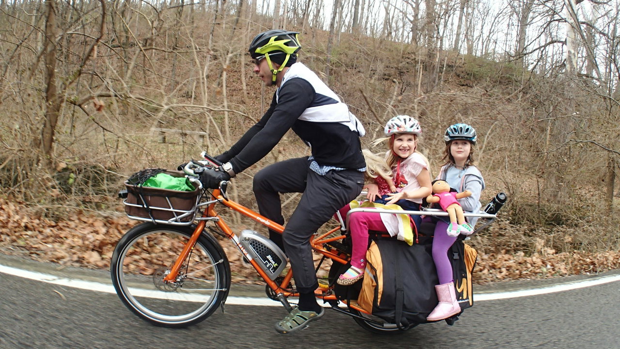 Mann mit Helm fährt ein orangefarbenes Longtail-Lastenrad, hinten sitzen zwei lachende Mädchen mit Fahrradhelmen