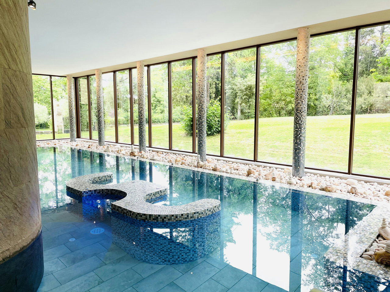 Hallenbad mit blauem Mosaikbecken, Säulen und raumhohen Fenstern zum grünen Garten, Kieselsteine am Beckenrand