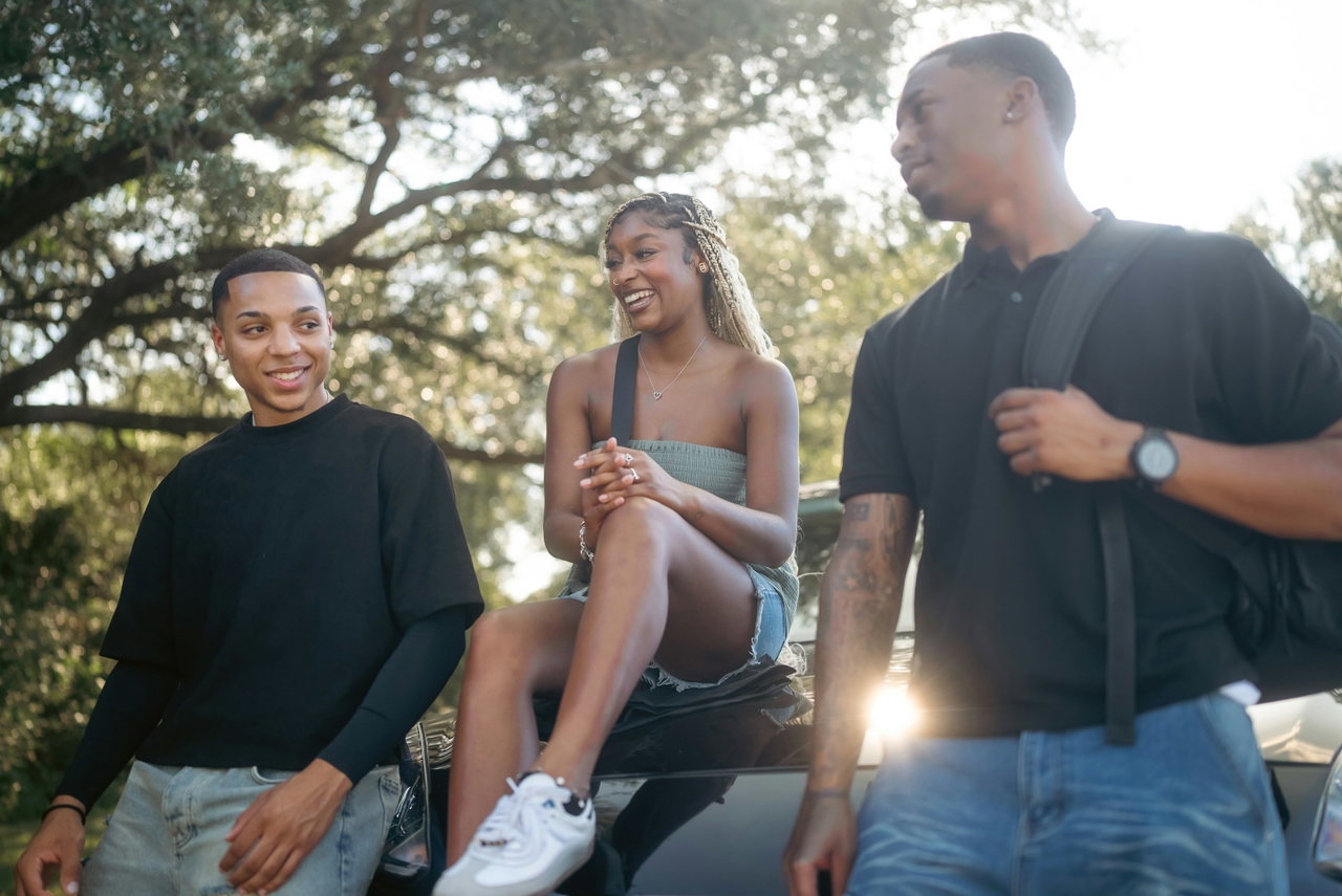 Eine junge Frau und zwei junge Männer lachen gemeinsam draußen unter Bäumen im warmen Sommerlicht