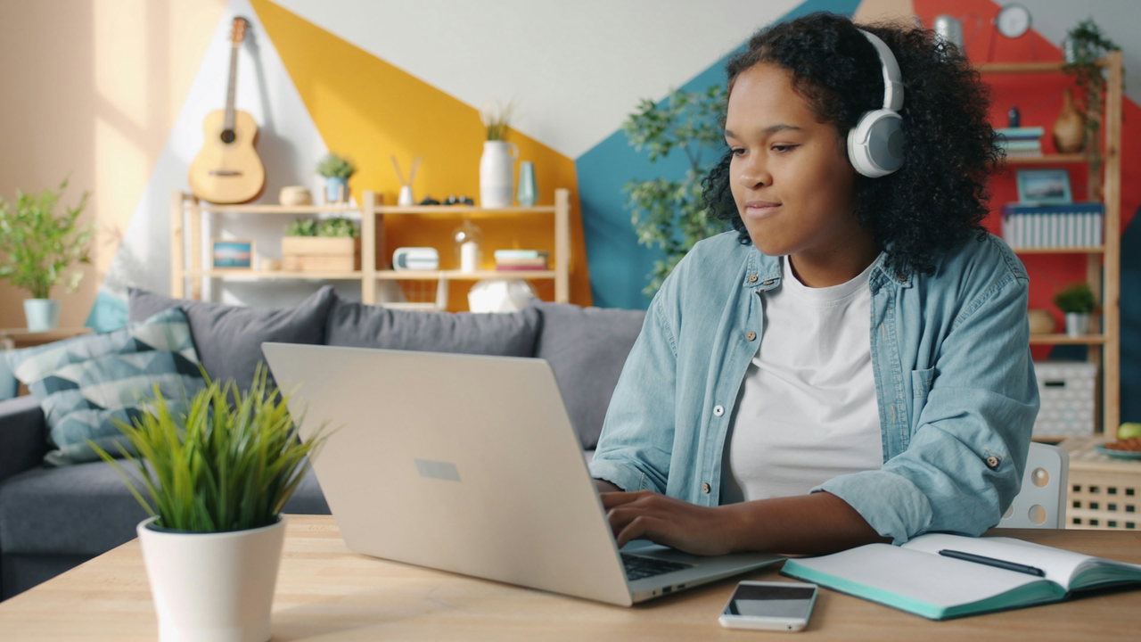 Jugendliche mit Kopfhörern sitzt zuhause am Laptop in einem bunten Zimmer mit Notizbuch und Gitarre