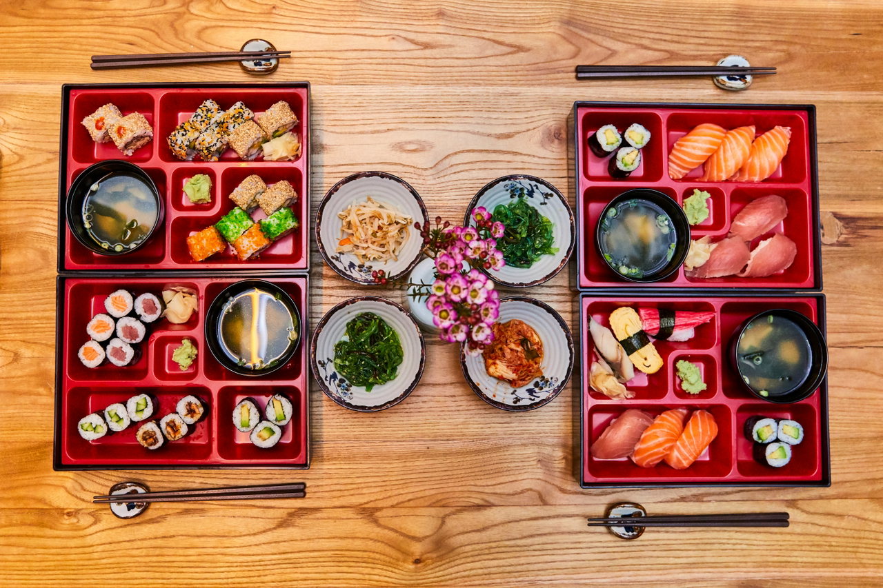 Vier rote Bento-Boxen mit Sushi, Misosuppe und kleinen Beilagen auf einem Holztisch, daneben Essstäbchen