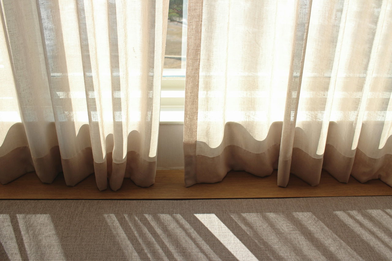 Helle Leinenvorhänge vor einem Fenster, durch die Sonne fällt Licht in Streifen auf Fensterbank und Boden.