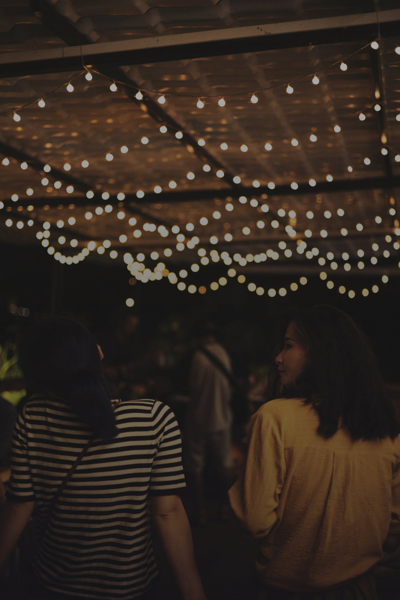 Zwei junge Frauen gehen abends unter warm leuchtenden Lichterketten, eine wendet den Kopf zur Seite