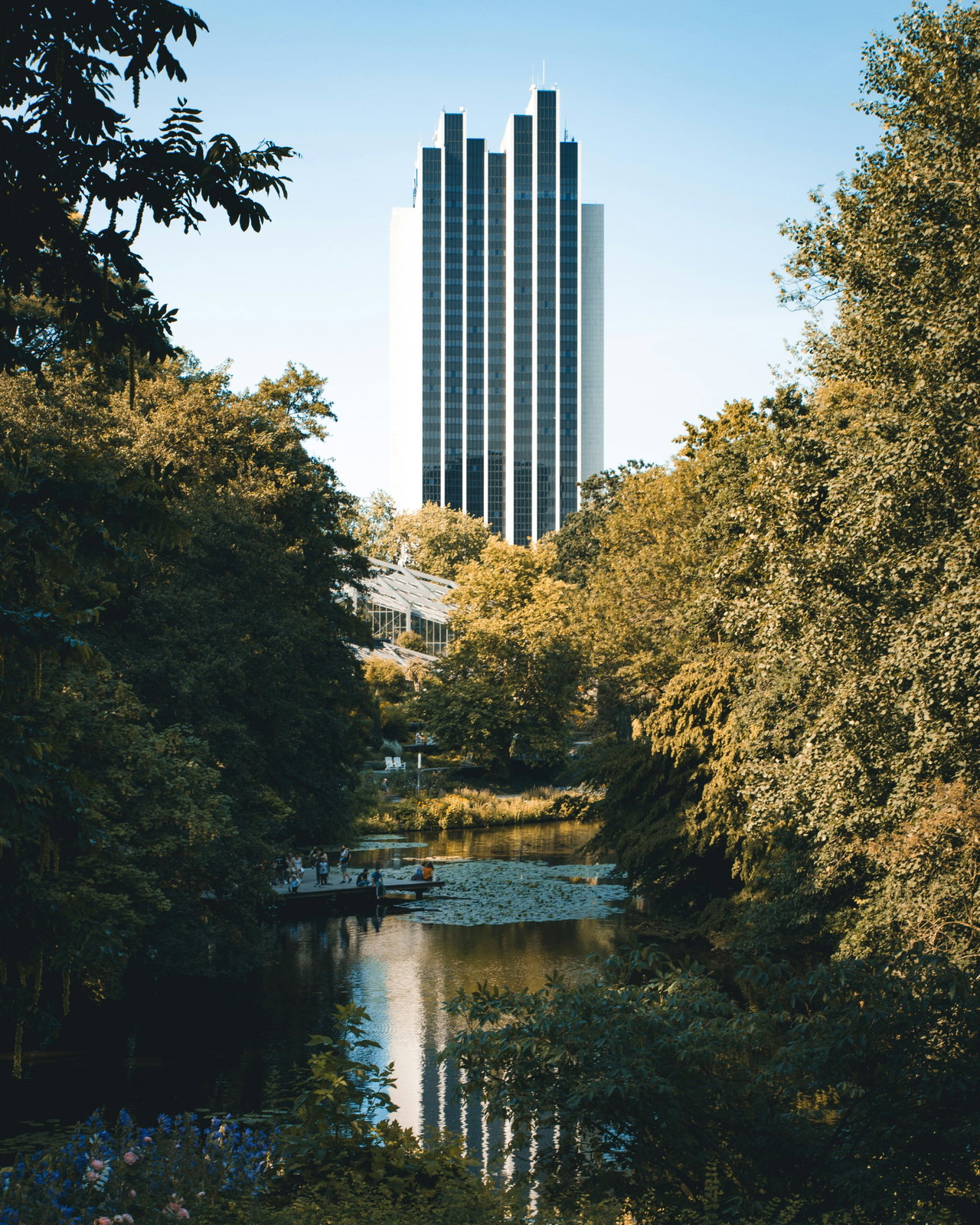 Teich zwischen hohen Bäumen, dahinter ein Hochhaus