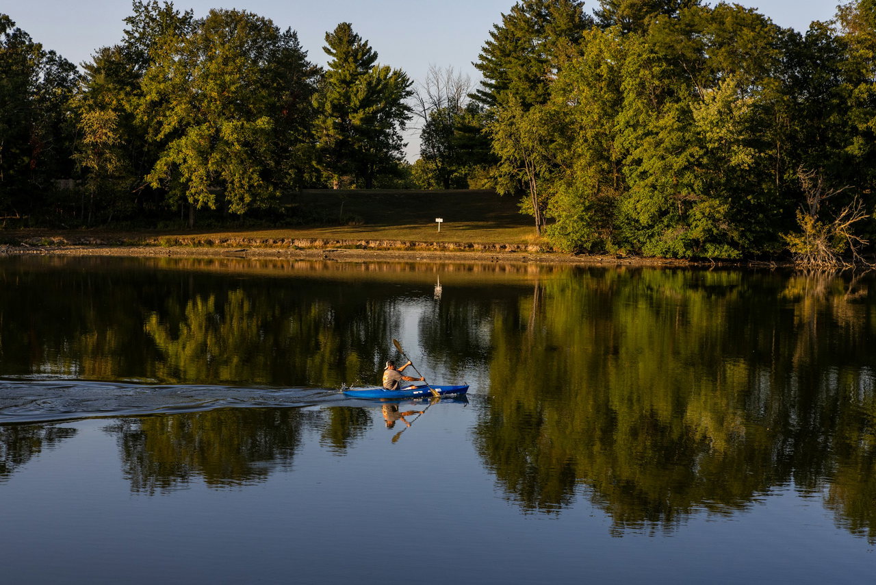 Ein Mann paddelt im blauen Kajak über ruhiges Wasser, in dem sich die Bäume am Ufer spiegeln.