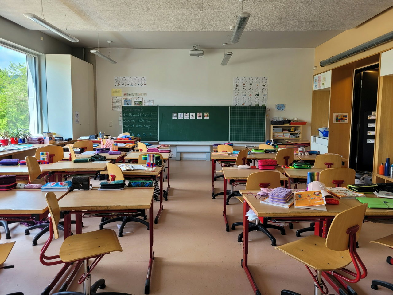 Helles Klassenzimmer mit Holztischen, Stühlen, Tafel und großen Fenstern mit Tageslicht