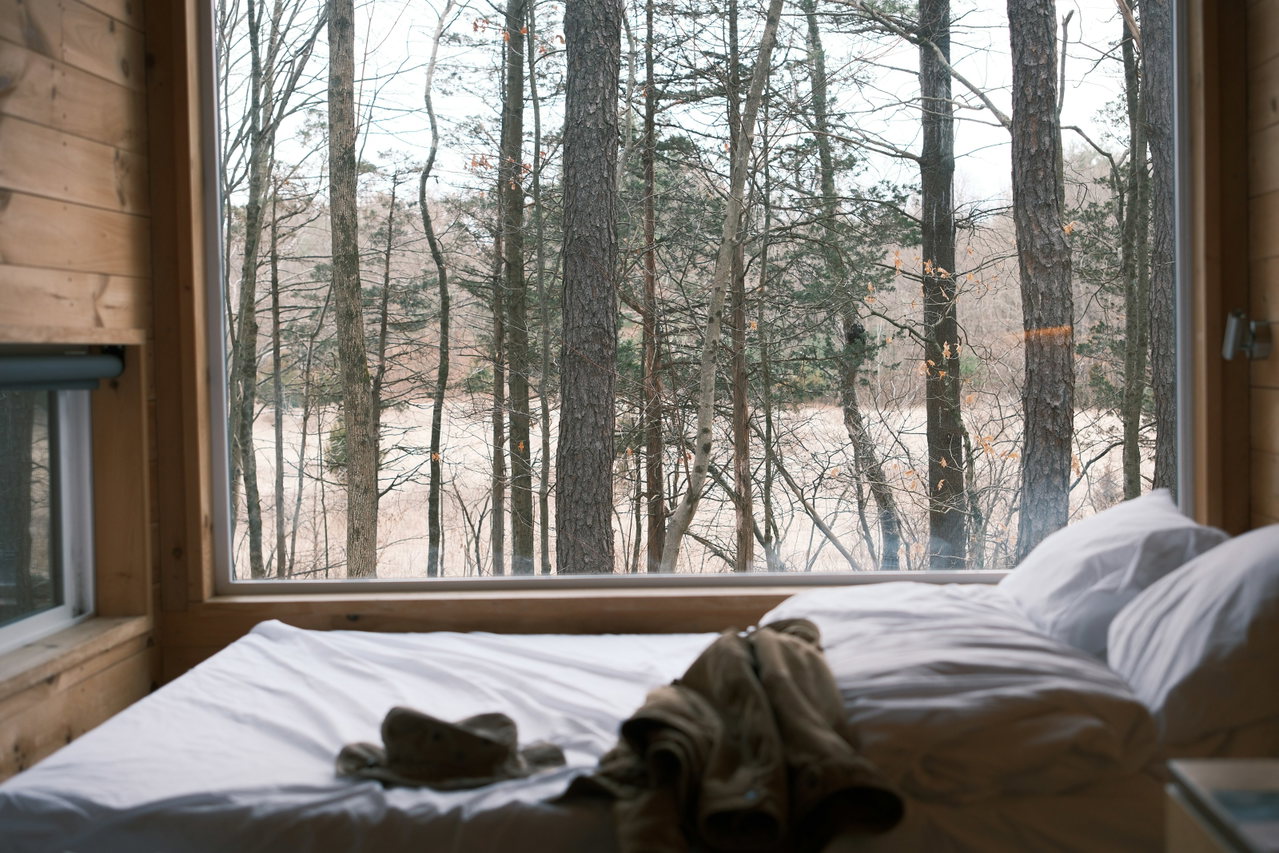 Holzzimmer mit großem Fenster und Blick auf kahle Waldbäume, Bett mit hellem Leinen
