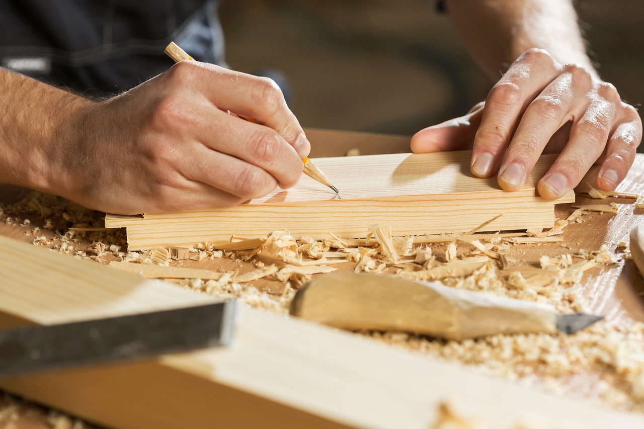 Hände eines Zimmermanns markieren mit einem Bleistift ein Holzbrett, daneben Hobelspäne