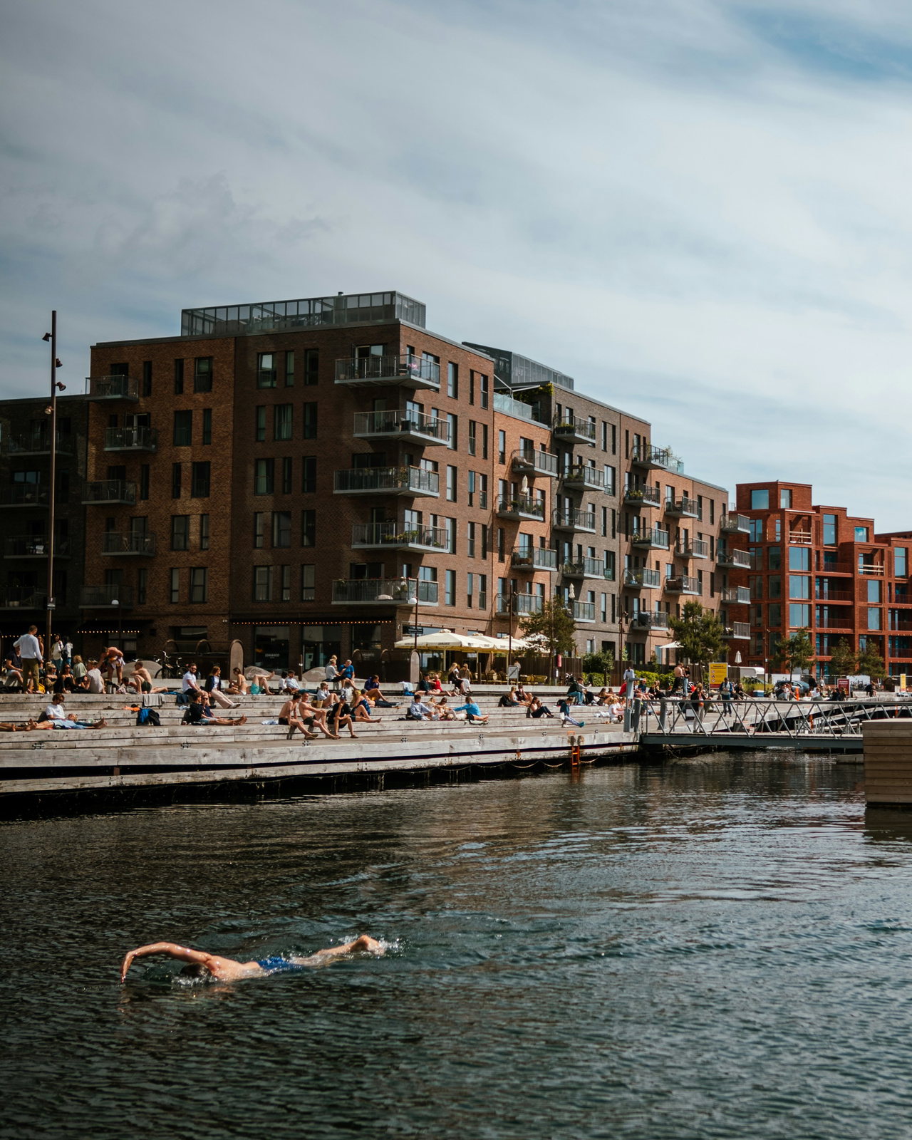 Wohnhäuser aus braunem und rotem Klinker mit Balkonen an einem Hafenbecken; auf den breiten Holzstufen am Kai sonnen sich viele Menschen, im Vordergrund schwimmt ein Mann.
