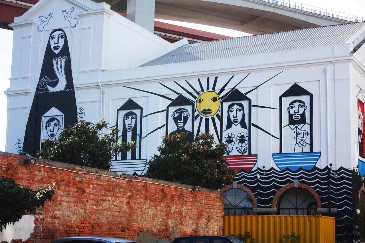 Großes Streetart-Wandbild mit Gesichtern und Sonnenstrahlen an einem weißen Gebäude unter einer Brücke in Lissabon