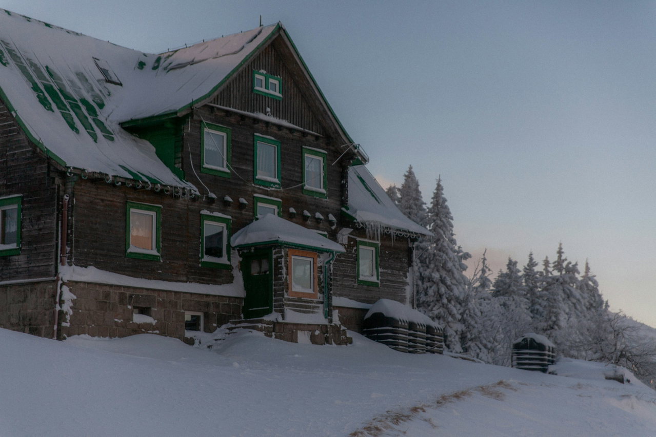 Dunkles Holzhaus mit grünem Dach im Abendlicht, tief verschneit am Rand eines Fichtenwalds
