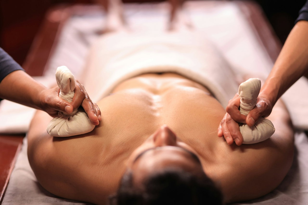 Zwei Hände legen warme Kräuterstempel auf den Oberkörper eines Gastes bei einer Massage
