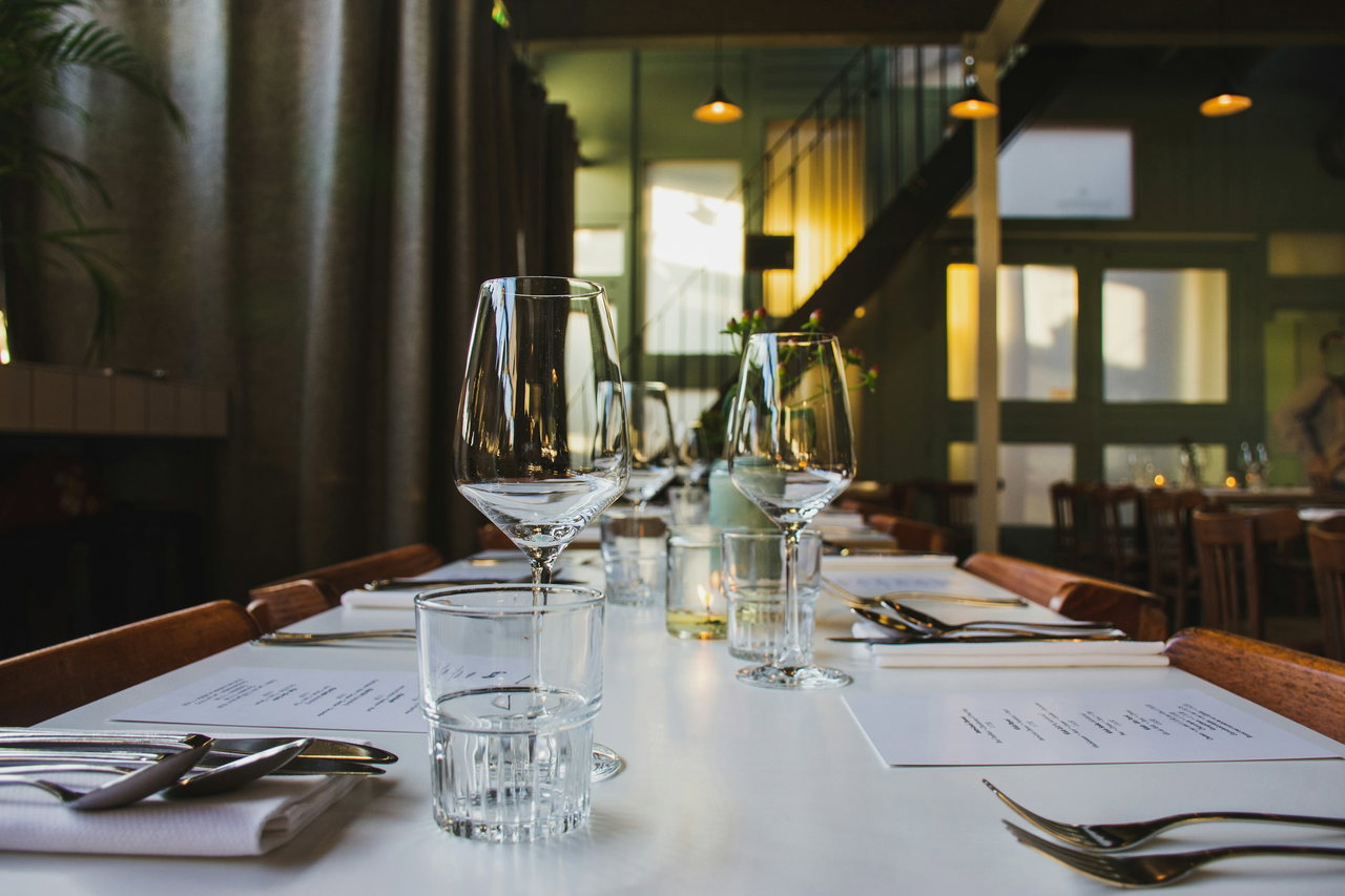 Festlich eingedeckte lange Tafel mit Weingläsern, Wassergläsern, Besteck und Menükarten im Restaurant mit Fenstertüren